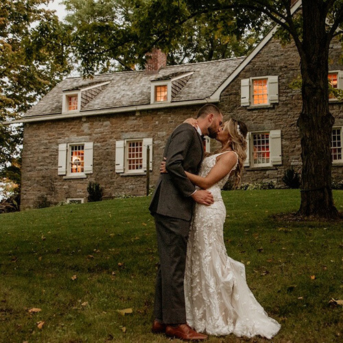 Married couple kissing in front of lit up brick house.